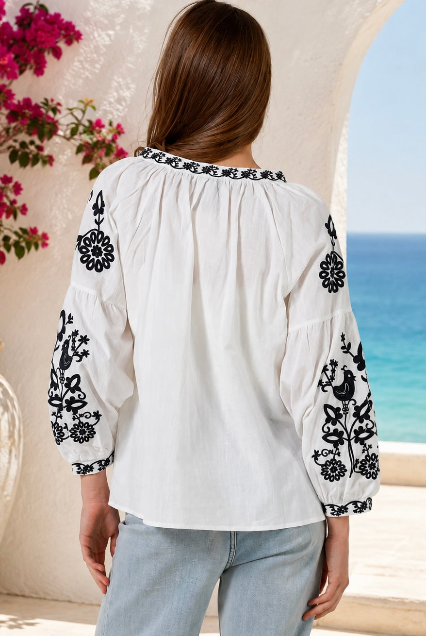 A woman viewed from behind wearing a white blouse with black floral embroidery on the sleeves and neckline, paired with light blue jeans, set against a Mediterranean-style background with a blue sea and white stucco walls.