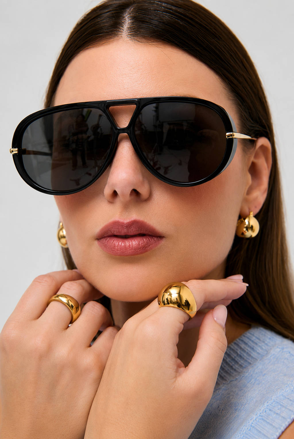A close-up of a woman wearing oversized black aviator-style sunglasses with gold metal temples, along with gold hoop earrings and gold rings, against a light gray background.