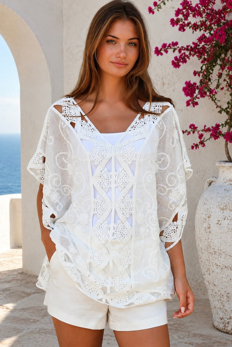 A woman wearing a white eyelet lace cover-up top with cutout details, standing outdoors in front of a white stucco wall with a blue ocean and pink flowers in the background.