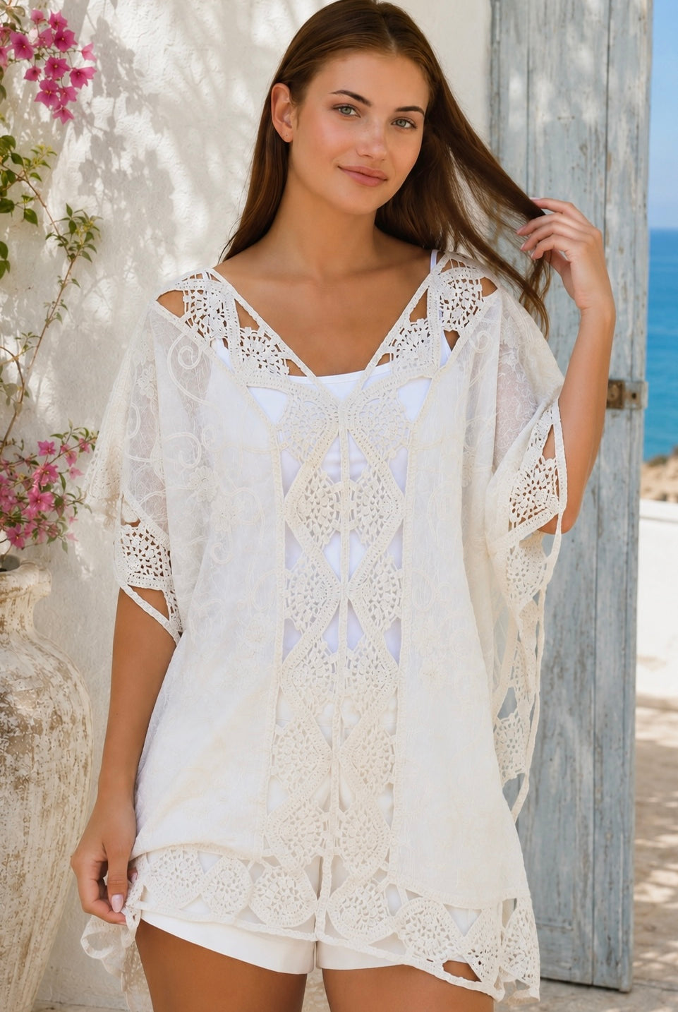 Cream crochet boho kaftan with ornate lace panels and relaxed sleeves in a sunlit Mediterranean coastal setting.