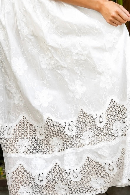 A woman wearing a white lace midi shirt dress with short puff sleeves, a tie waist, and a front slit, standing outdoors in front of tropical green foliage.