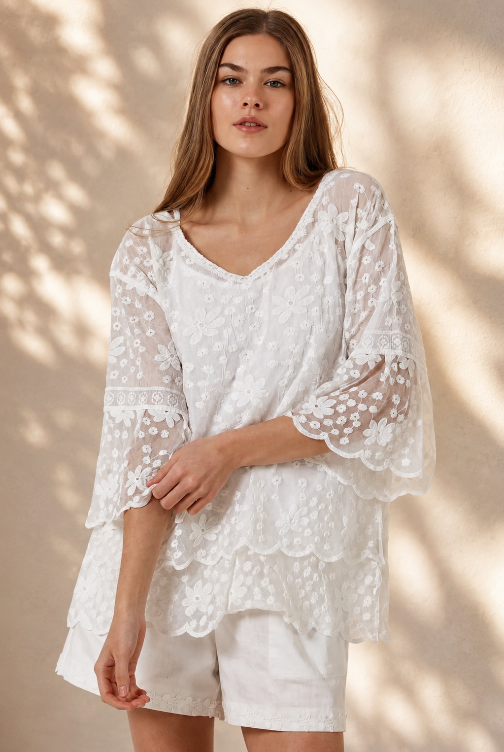A woman wearing a white lace long-sleeve tunic top with matching white shorts, standing in a sunlit outdoor setting with a beige wall and potted plant in the background.