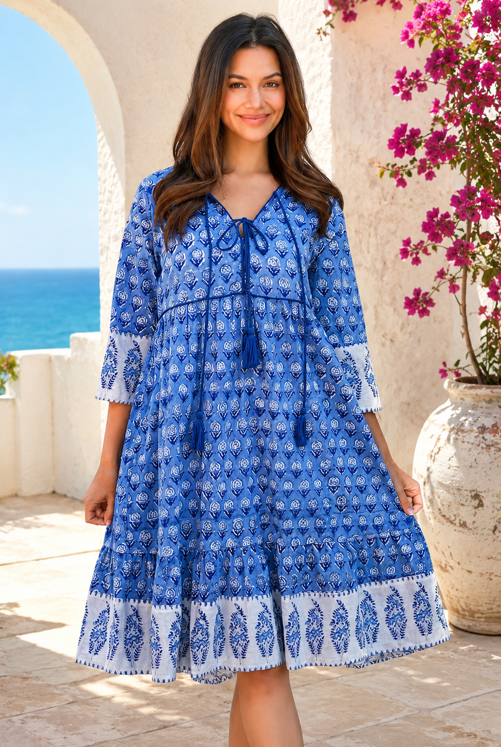 A woman wearing a blue and white patterned bohemian-style dress, standing in front of a Mediterranean-style stone wall with a view of the blue sea in the background from Bohemian Love Runway