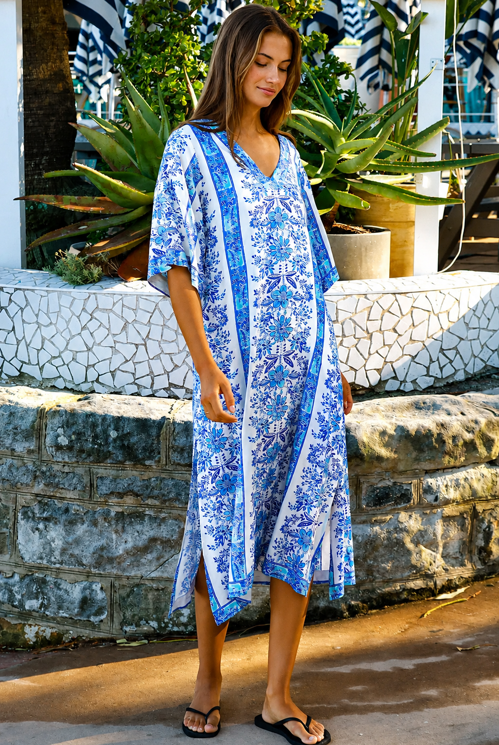 A woman wearing a blue and white patterned kaftan dress with a V-neck and three-quarter sleeves, walking outdoors in front of a stone wall and tropical plants.