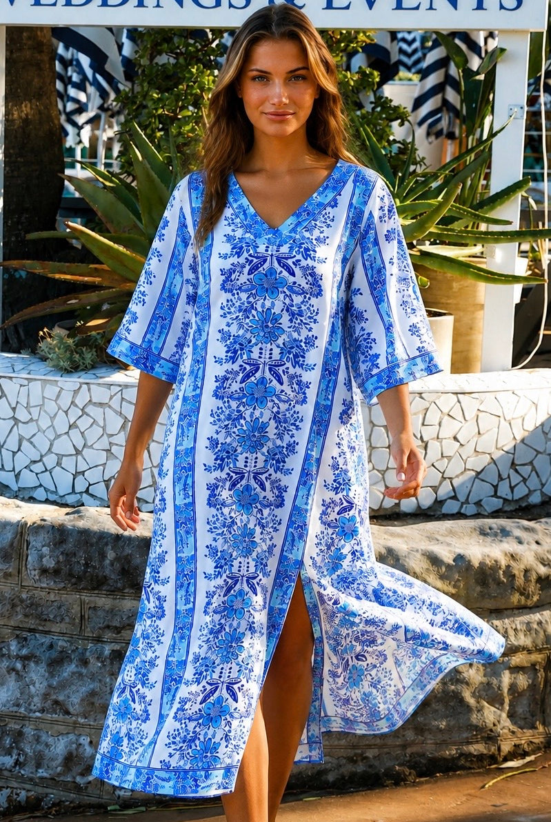 A woman wearing a white kaftan-style tunic dress with a vibrant blue floral and geometric pattern, photographed outdoors with green foliage in the background.