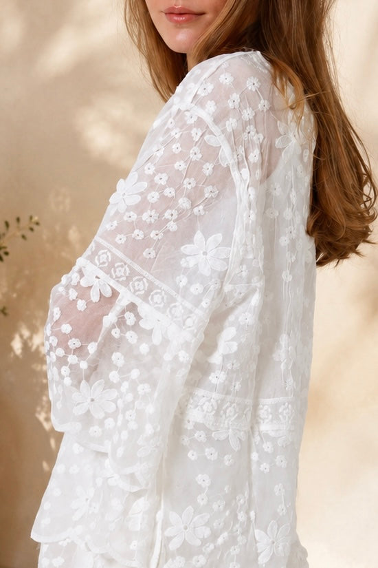 A woman wearing a white lace long-sleeve tunic top with matching white shorts, standing in a sunlit outdoor setting with a beige wall and potted plant in the background.