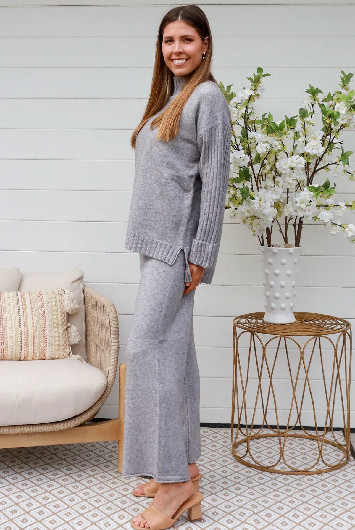 Woman wearing grey knit lounge set with high-neck jumper and wide-leg pants standing in front of a white timber wall - Mellow Lounge Set Grey Mist Bohemian Love Runway