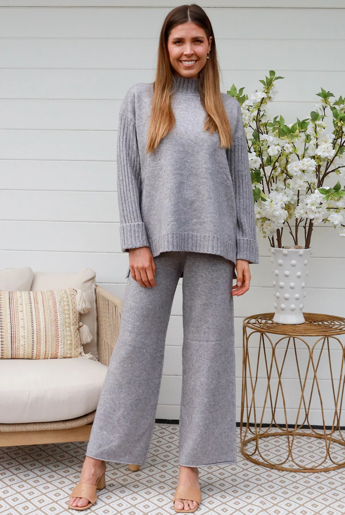 Woman wearing grey knit lounge set with high-neck jumper and wide-leg pants standing in front of a white timber wall - Mellow Lounge Set Grey Mist Bohemian Love Runway