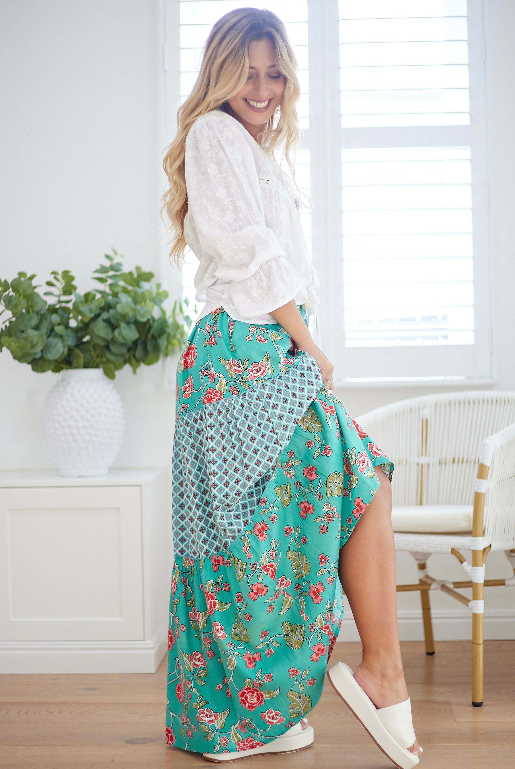 Woman wearing a white blouse and a long turquoise floral skirt in a bright room.