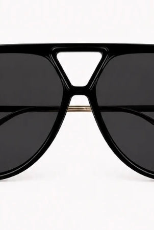 A close-up of a woman wearing oversized black aviator-style sunglasses with gold metal temples, along with gold hoop earrings and gold rings, against a light gray background.