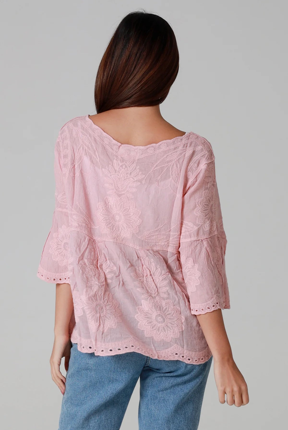 Woman wearing a blush pink embroidered cotton blouse with scalloped neckline and flared sleeves, styled with denim jeans - romantic boho top Australia.