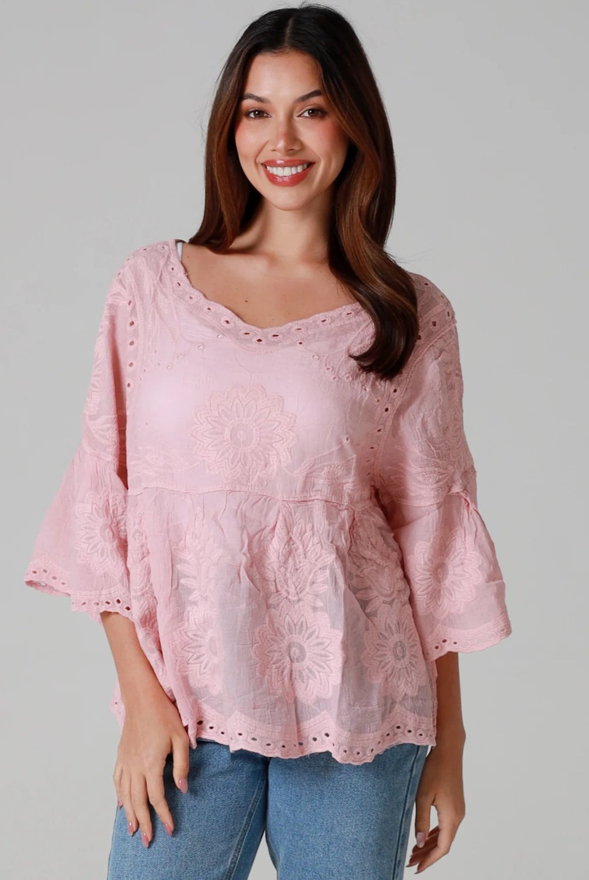 Woman wearing a blush pink embroidered cotton blouse with scalloped neckline and flared sleeves, styled with denim jeans - romantic boho top Australia.