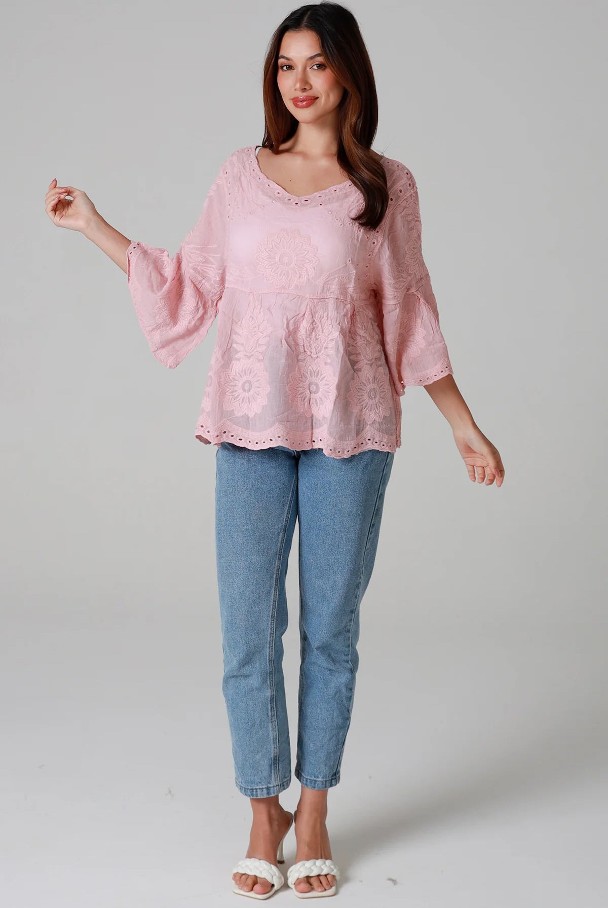 Woman wearing a blush pink embroidered cotton blouse with scalloped neckline and flared sleeves, styled with denim jeans - romantic boho top Australia.