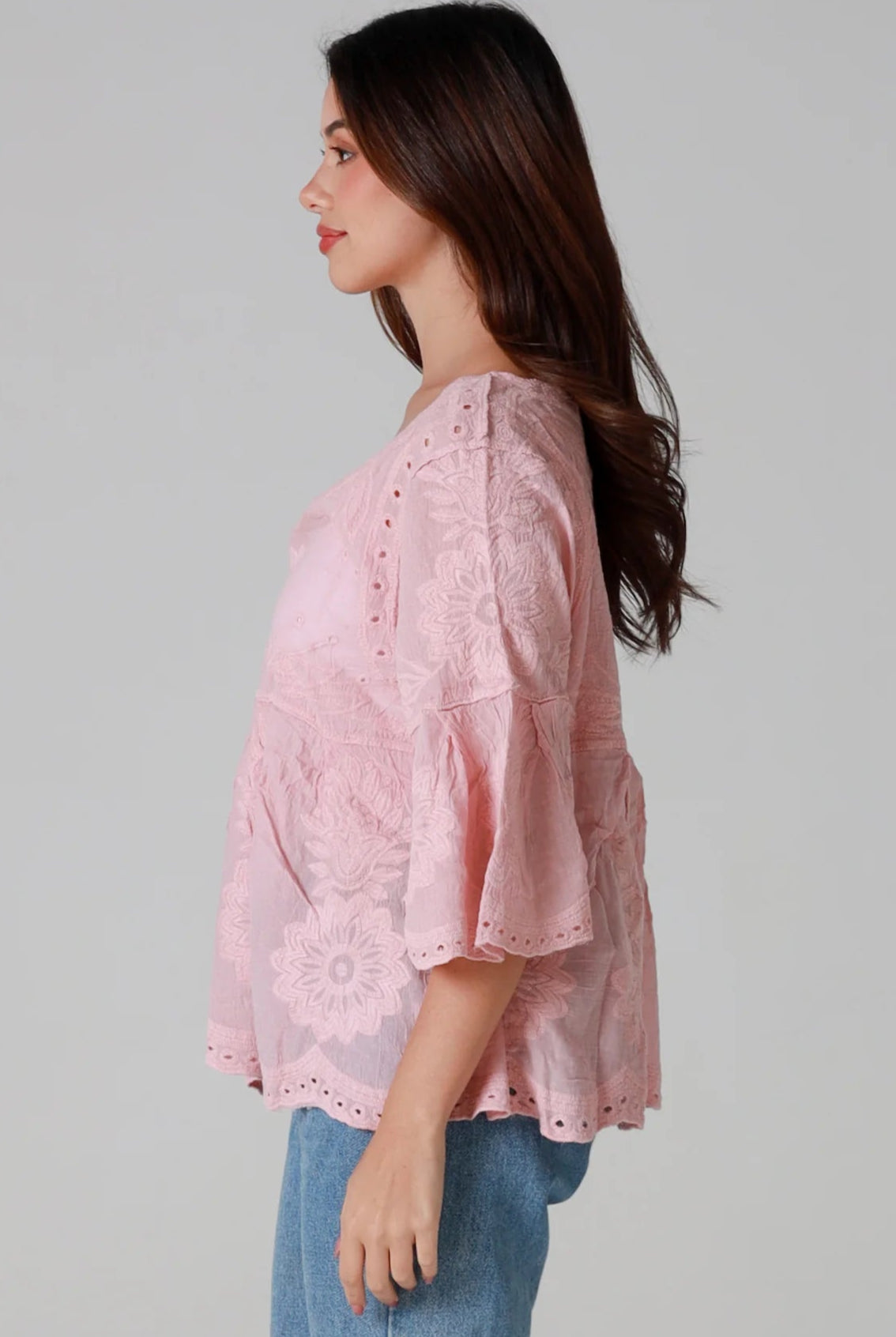 Woman wearing a blush pink embroidered cotton blouse with scalloped neckline and flared sleeves, styled with denim jeans - romantic boho top Australia.