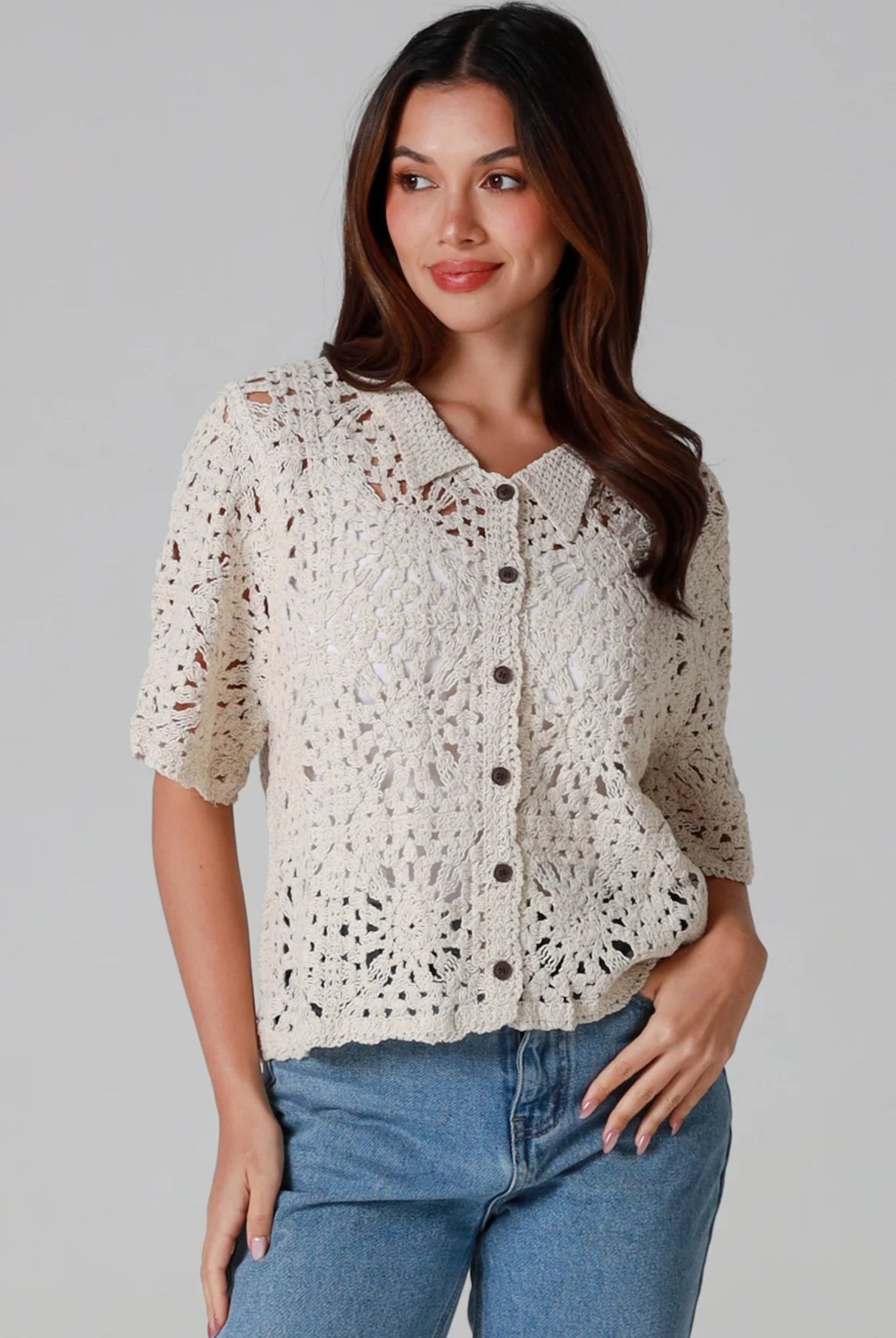 Woman wearing a beige short-sleeve crochet blouse with scalloped collar and buttons, styled open over jeans - boho cardigan top Australia.