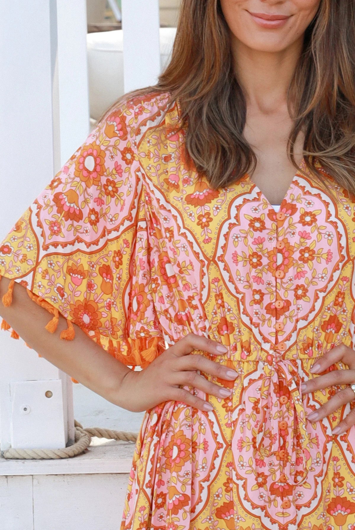 Woman wearing a vintage-inspired boho maxi dress in warm coral and apricot floral print, featuring flutter sleeves, a drawstring waist, and a flowing tiered skirt, styled outdoor in soft summer light.