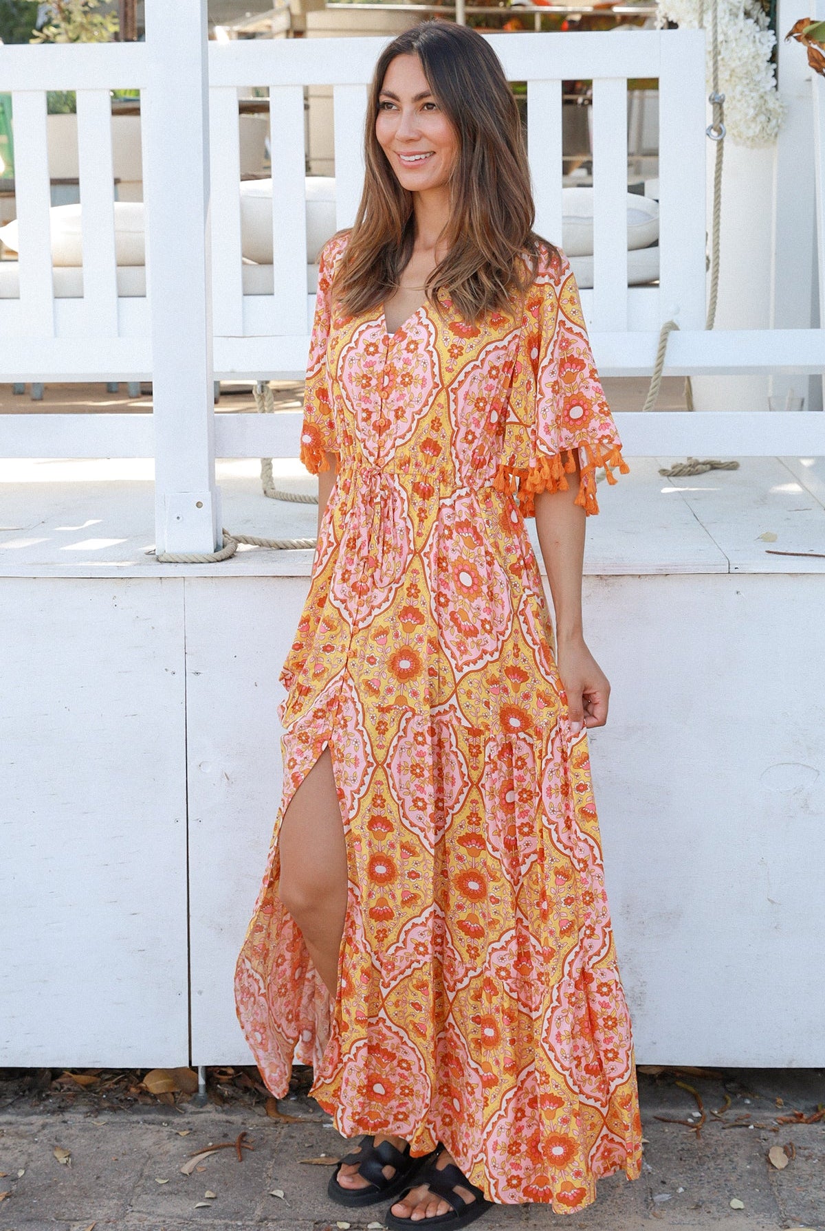 Woman wearing a vintage-inspired boho maxi dress in warm coral and apricot floral print, featuring flutter sleeves, a drawstring waist, and a flowing tiered skirt, styled outdoor in soft summer light.