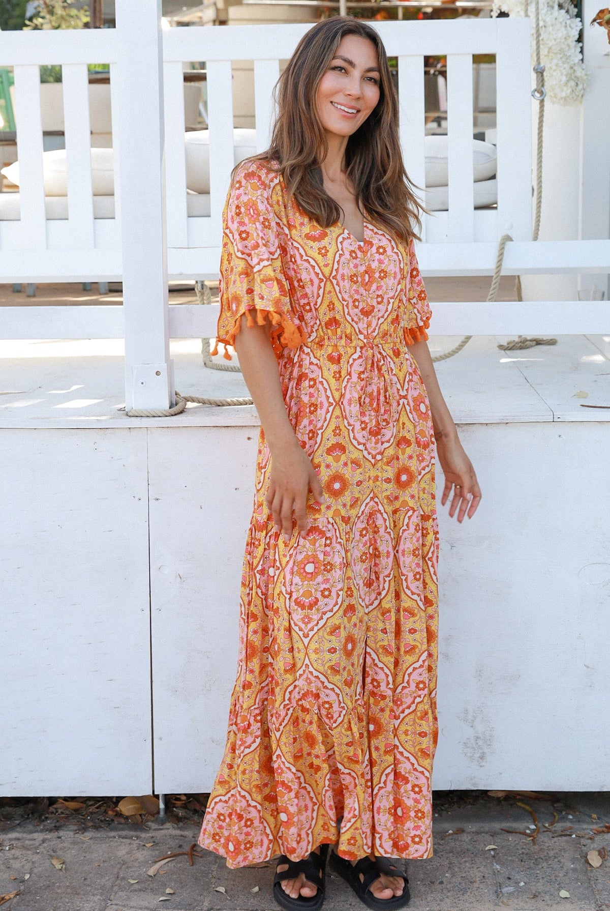 Woman wearing a vintage-inspired boho maxi dress in warm coral and apricot floral print, featuring flutter sleeves, a drawstring waist, and a flowing tiered skirt, styled outdoor in soft summer light.