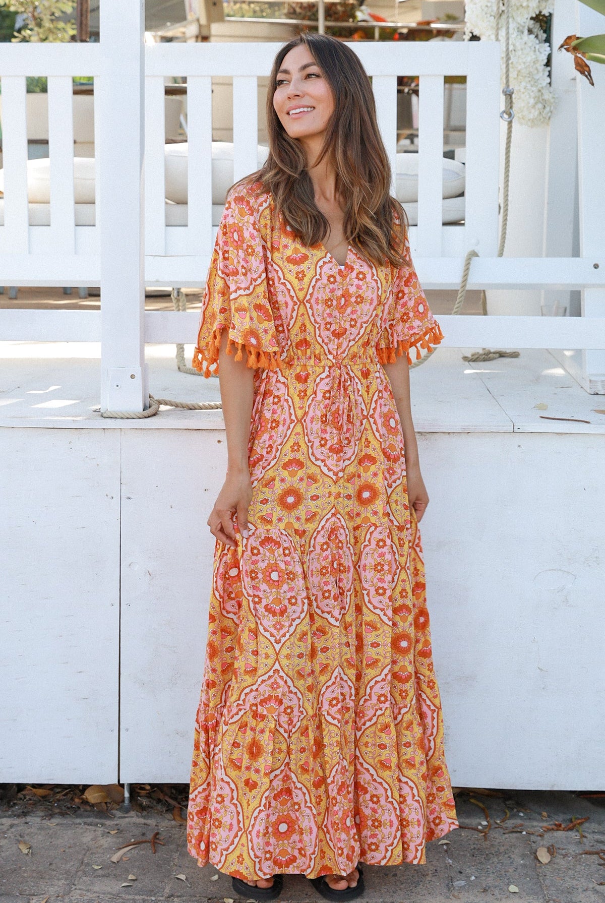 Woman wearing a vintage-inspired boho maxi dress in warm coral and apricot floral print, featuring flutter sleeves, a drawstring waist, and a flowing tiered skirt, styled outdoor in soft summer light.