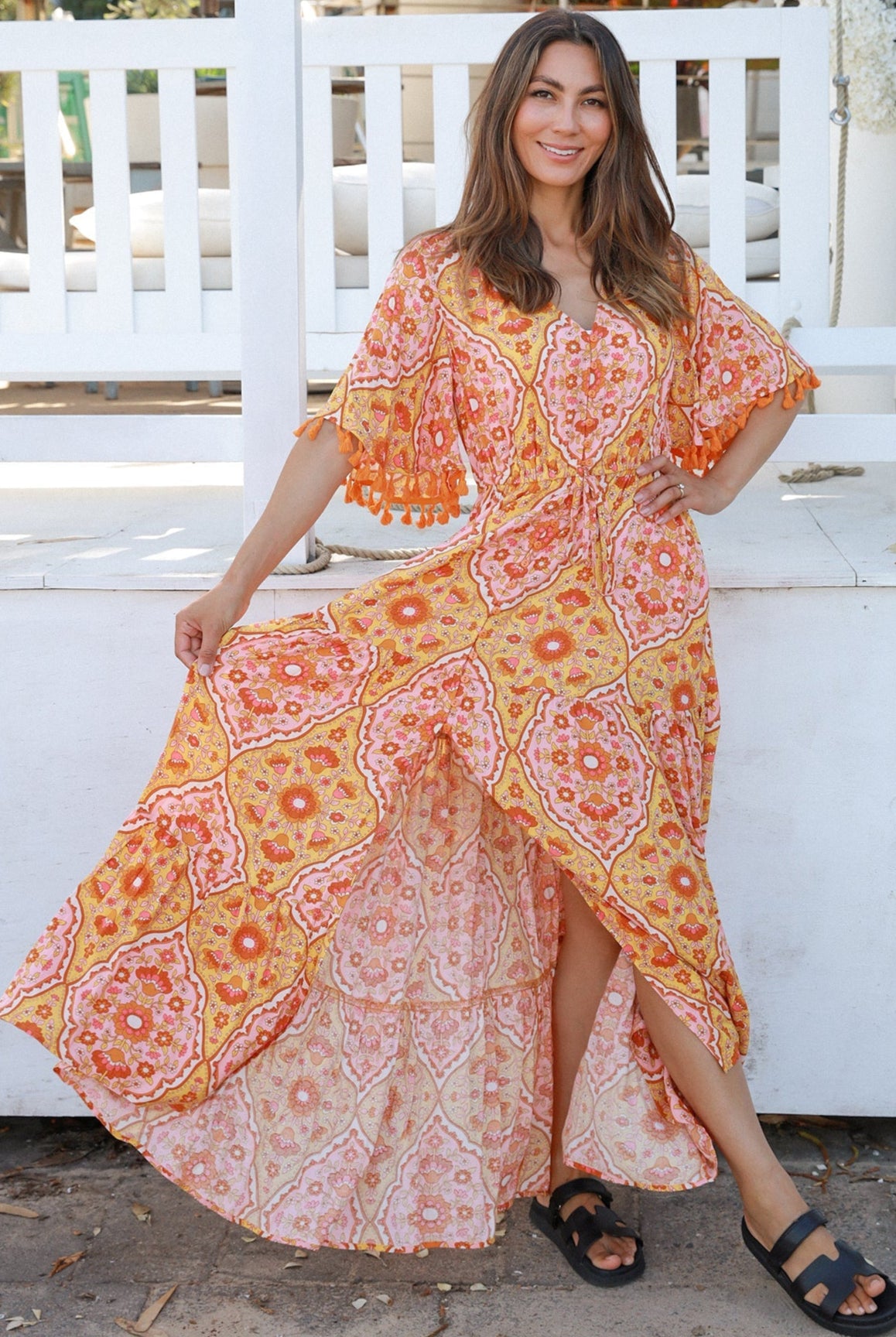 Woman wearing a vintage-inspired boho maxi dress in warm coral and apricot floral print, featuring flutter sleeves, a drawstring waist, and a flowing tiered skirt, styled outdoor in soft summer light.