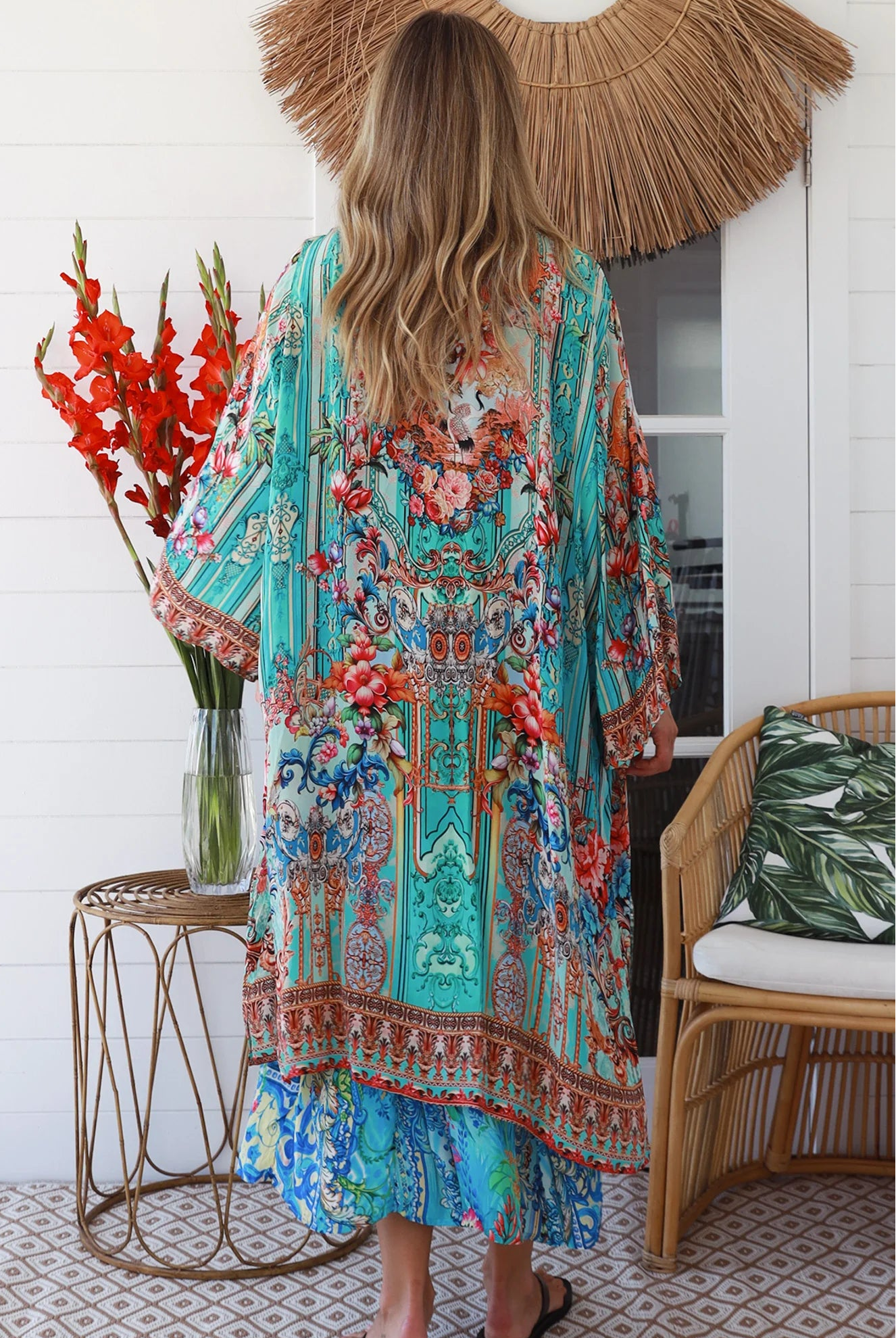 Woman in turquoise floral crepe - viscose kimono with bronze and sapphire border detailing, walking along a sunlit coastal setting - resort luxe boho style.