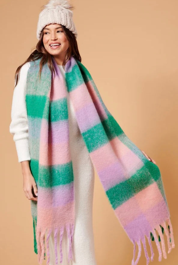 Woman wearing a chunky white knit beanie and oversized fluffy scarf in pastel green, pink and lilac stripes, styled with a neutral winter outfit.