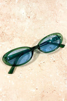 Green retro oval sunglasses with blue tinted lenses styled on stone background - vintage flat lay eyewear Australia.