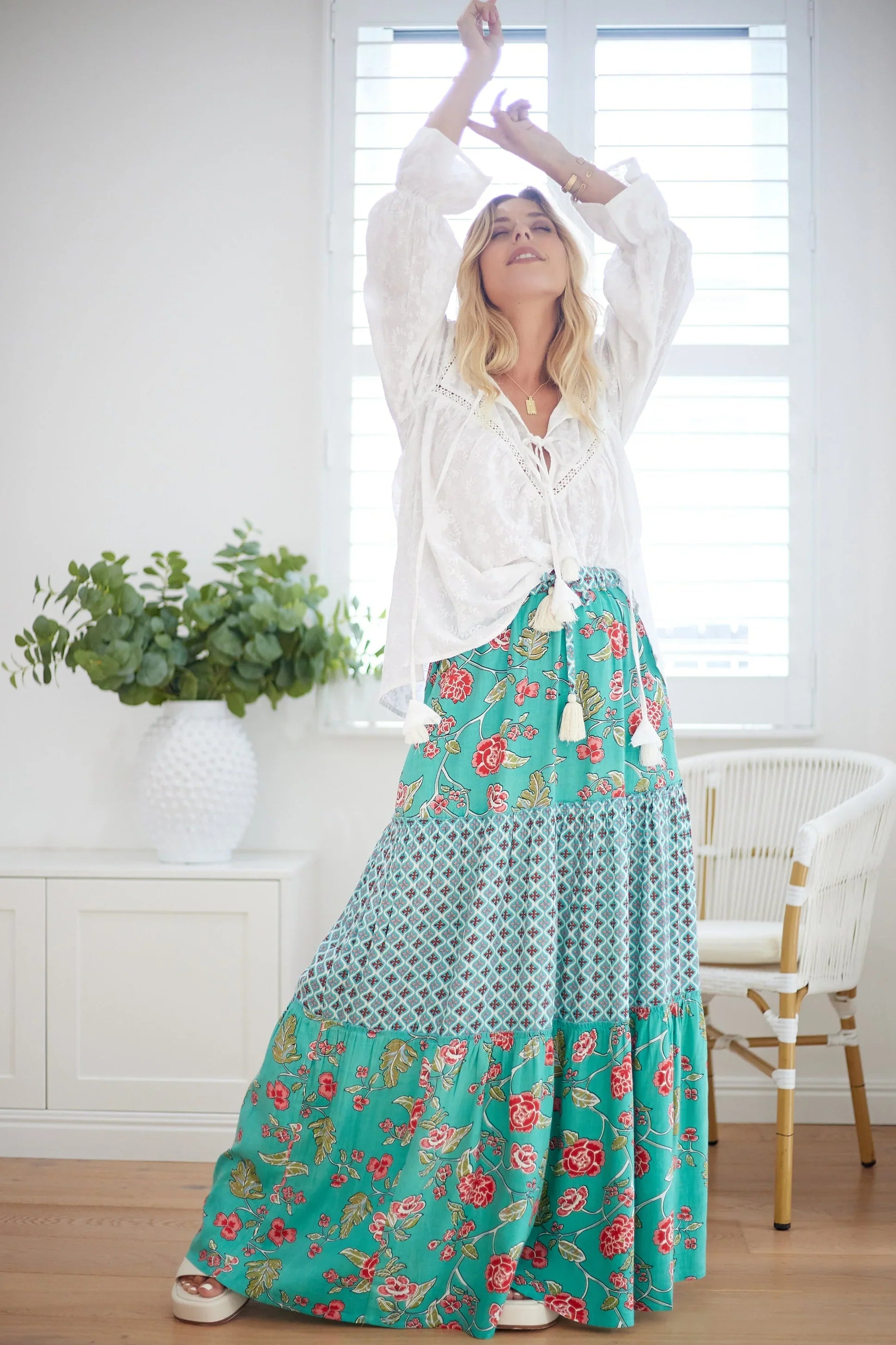 Woman wearing a white boho blouse and green floral tiered maxi skirt, standing barefoot indoors with arms raised; a free-spirited bohemian fashion look