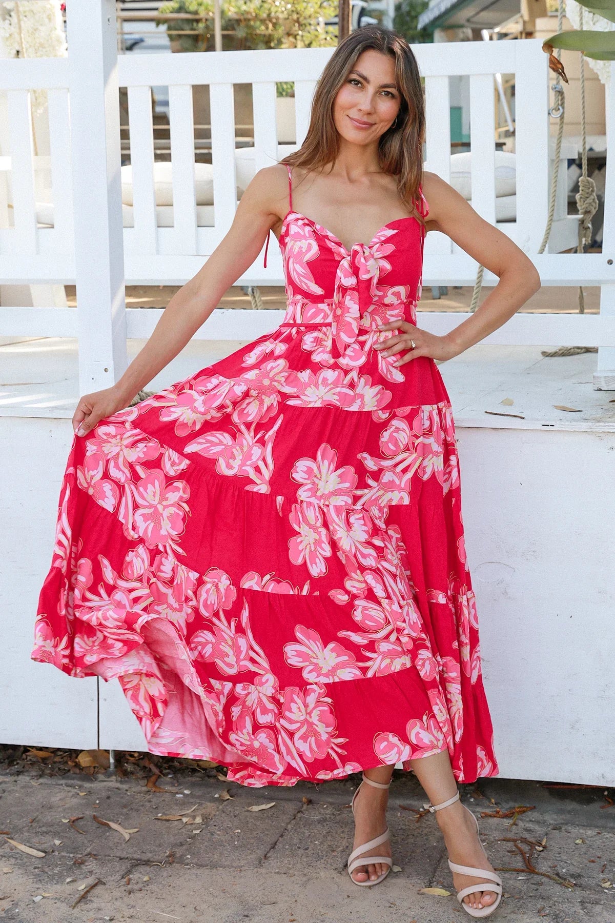 Woman wearing a vibrant red and pink foral bohemian maxi dress, smiling and lifting the hem slightly standing in front of a white fence.