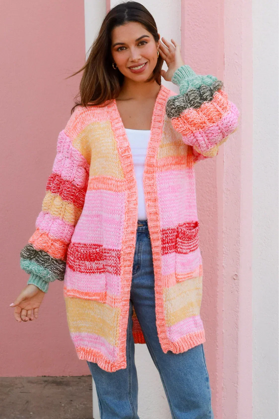 Smiling woman in rainbow patch knit cardigan and jeans in front of pink wall - colourful boho knitwear Australia.