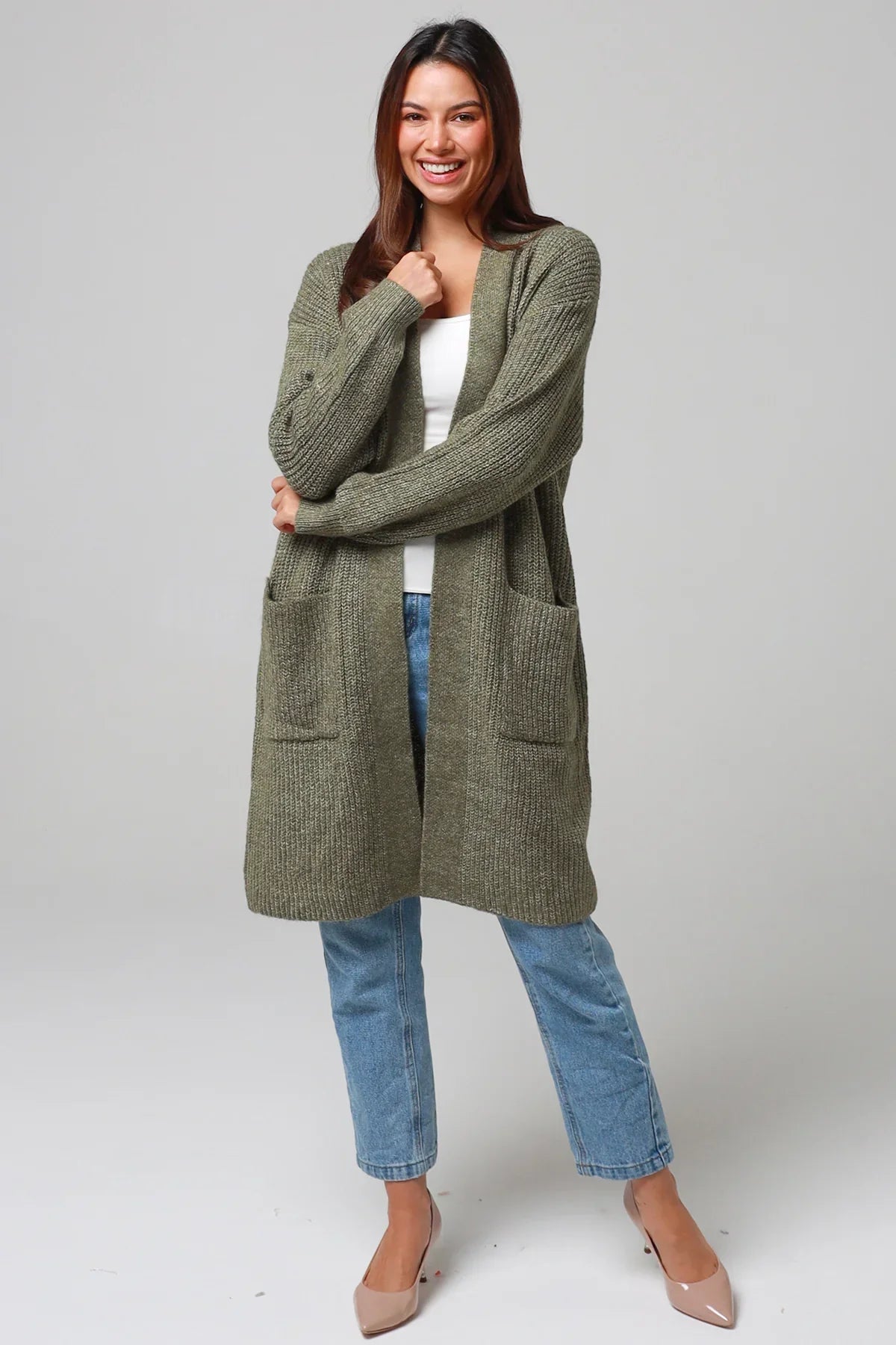 Woman wearing an olive green longline cardigan over a white top and denim jeans, smiling with arms folded— cozy bohemian winter knitwear style.