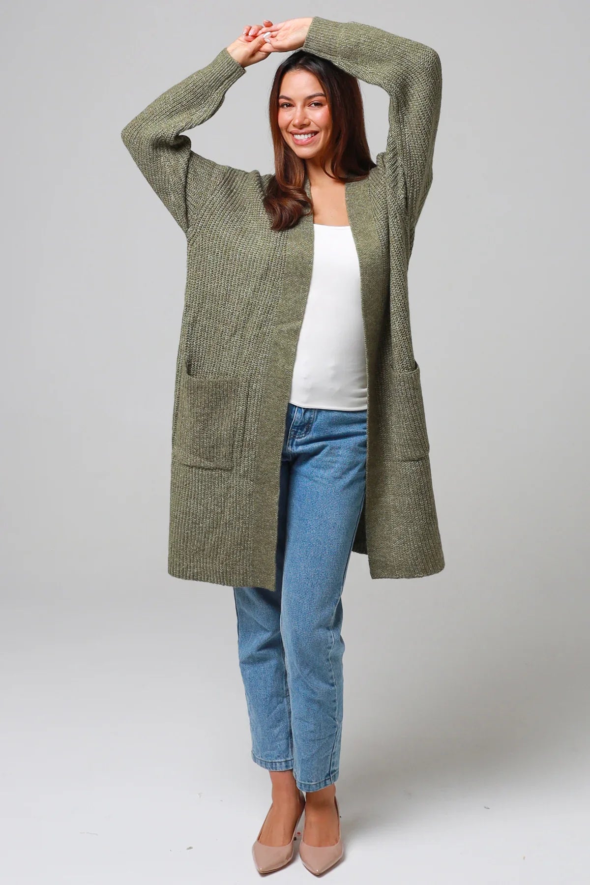 Woman wearing a long sage green knit cardigan over a white top and blue jeans, smiling with arms raised - cozy boho winter style Australia.
