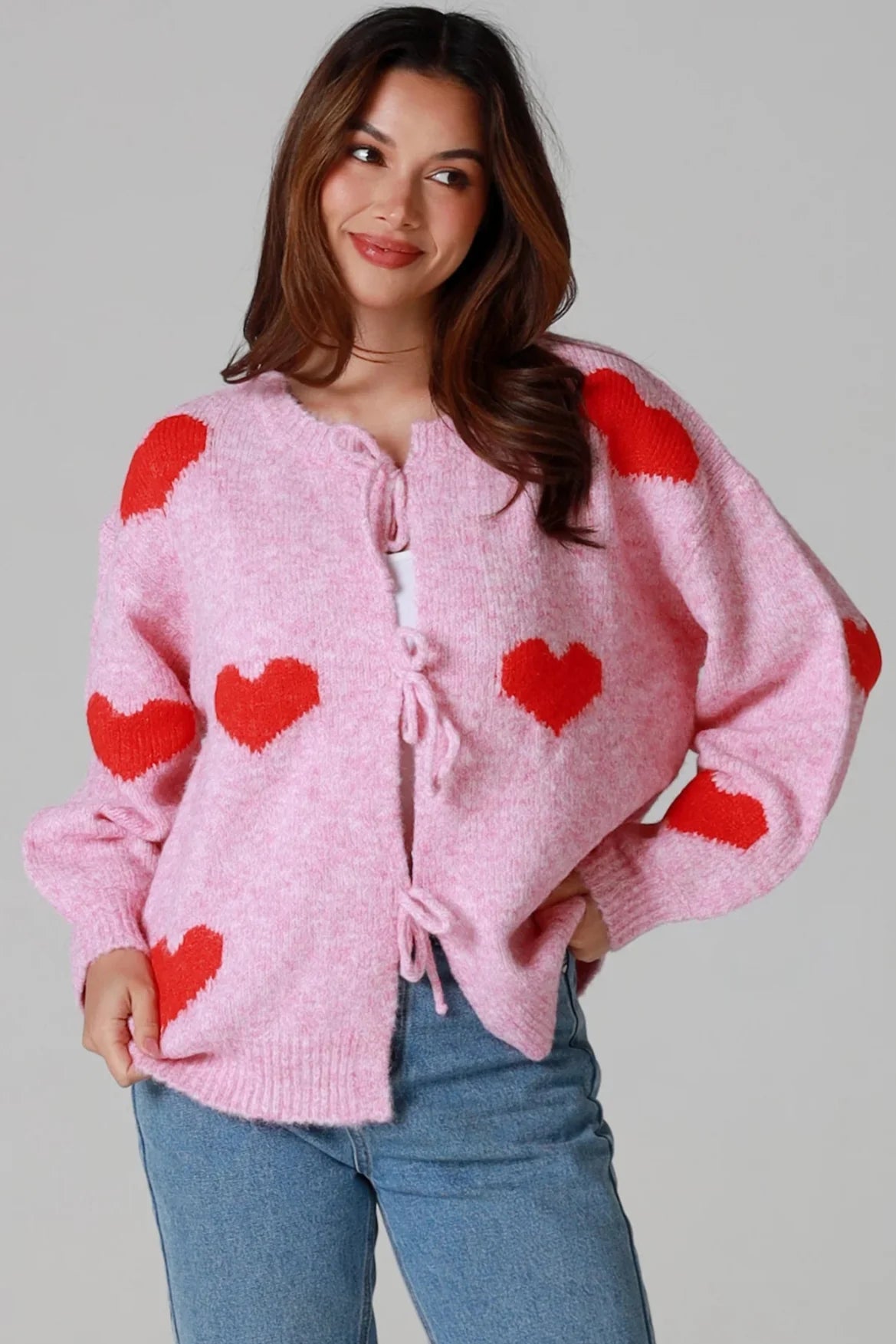 Woman wearing pink knit cardigan with red heart print and blue jeans smiling softly - Boho knitwear Australia.