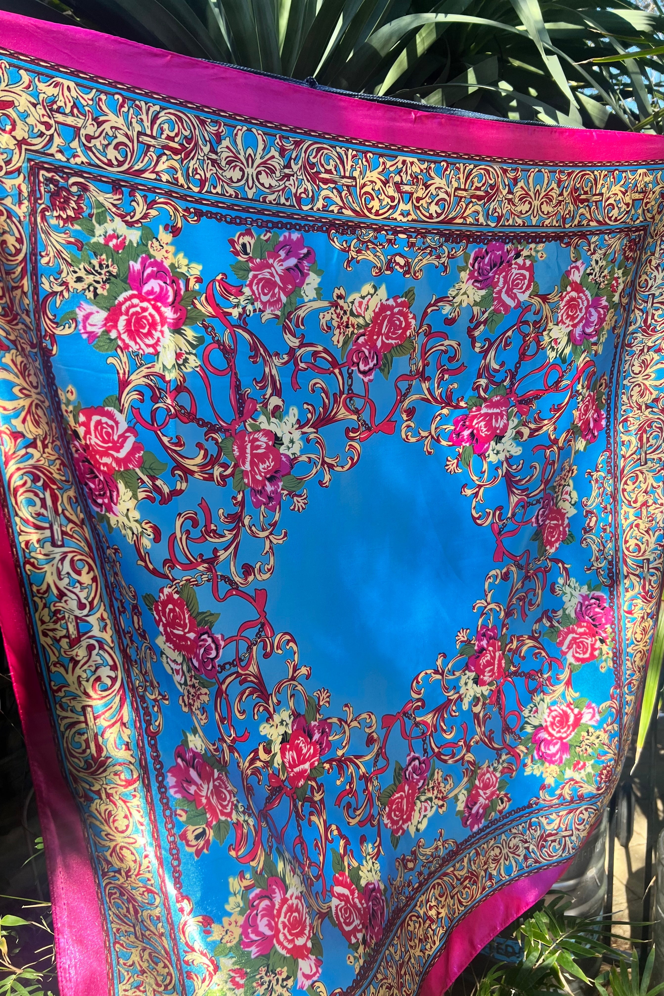 Boho gypsy-style scarf with bright blue centre, magenta border, and intricate floral print in pink gold, and green — displayed outdoors against foliage.