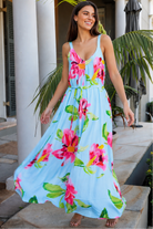 A woman wearing a light blue sleeveless maxi dress with a vibrant pink and green floral print, standing on stone steps in front of a building with white columns and tropical plants.