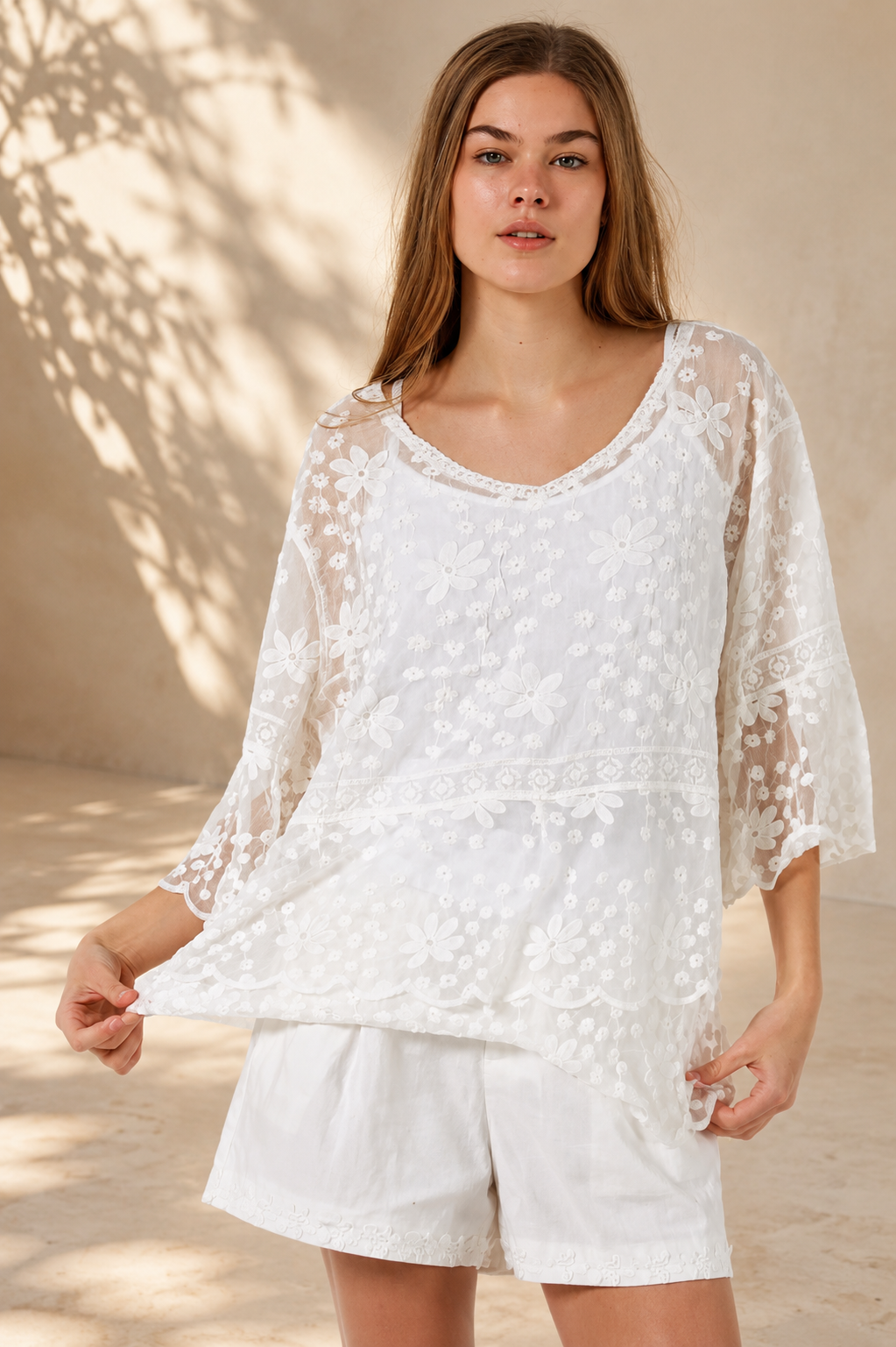 A woman wearing a white lace long-sleeve tunic top with matching white shorts, standing in a sunlit outdoor setting with a beige wall and potted plant in the background.