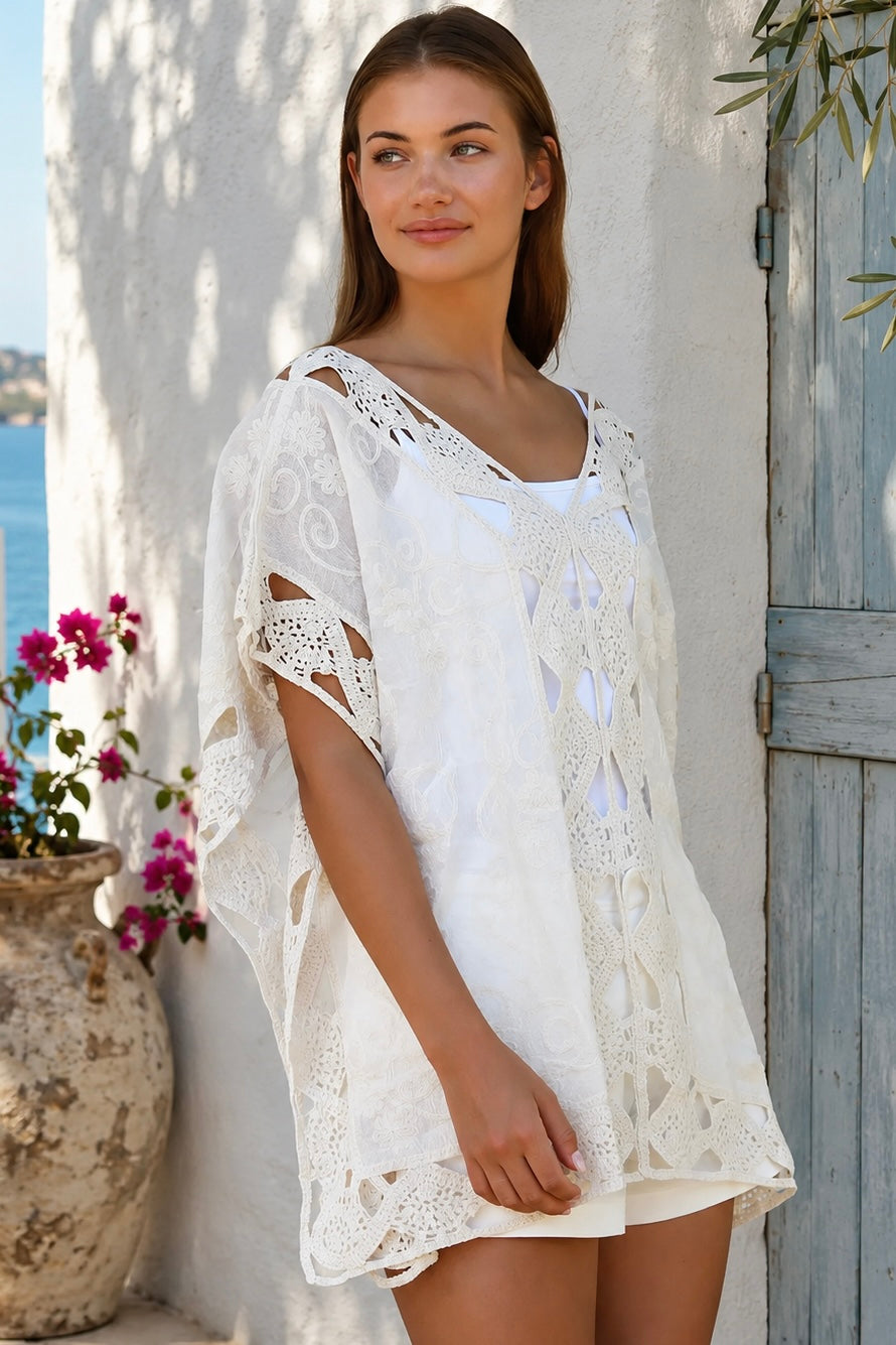 Cream crochet boho kaftan with ornate lace panels and relaxed sleeves in a sunlit Mediterranean coastal setting.