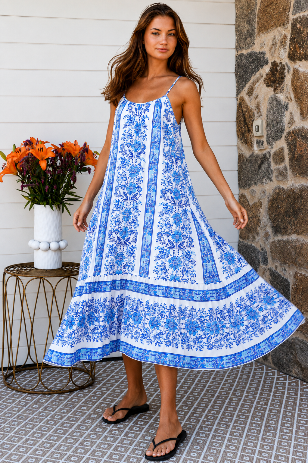 A woman modeling a blue and white patterned maxi dress with spaghetti straps, standing on a patterned tile porch next to a small table with orange flowers.