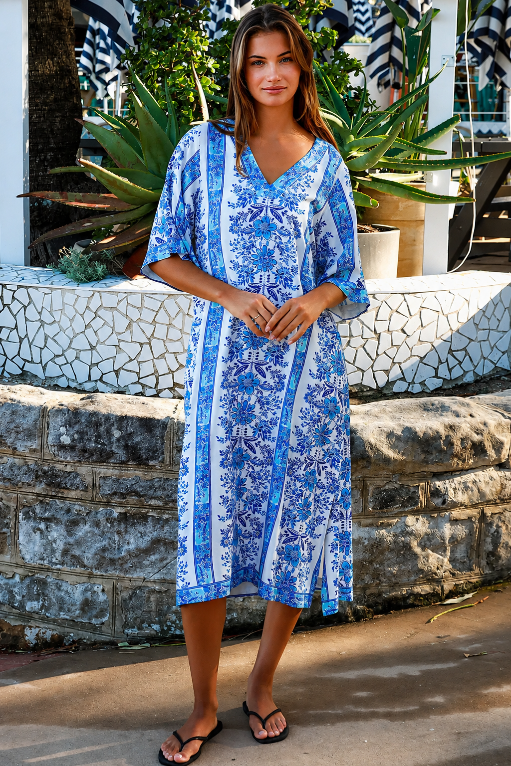 A woman wearing a blue and white patterned kaftan dress with a V-neck and three-quarter sleeves, walking outdoors in front of a stone wall and tropical plants.