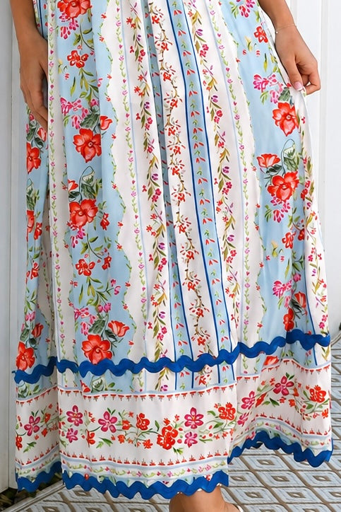 A woman wearing a sleeveless, A-line maxi dress with a white base, blue scalloped trim, and a red and pink floral print, standing outdoors in front of green plants.