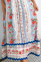 A woman wearing a sleeveless, A-line maxi dress with a white base, blue scalloped trim, and a red and pink floral print, standing outdoors in front of green plants.