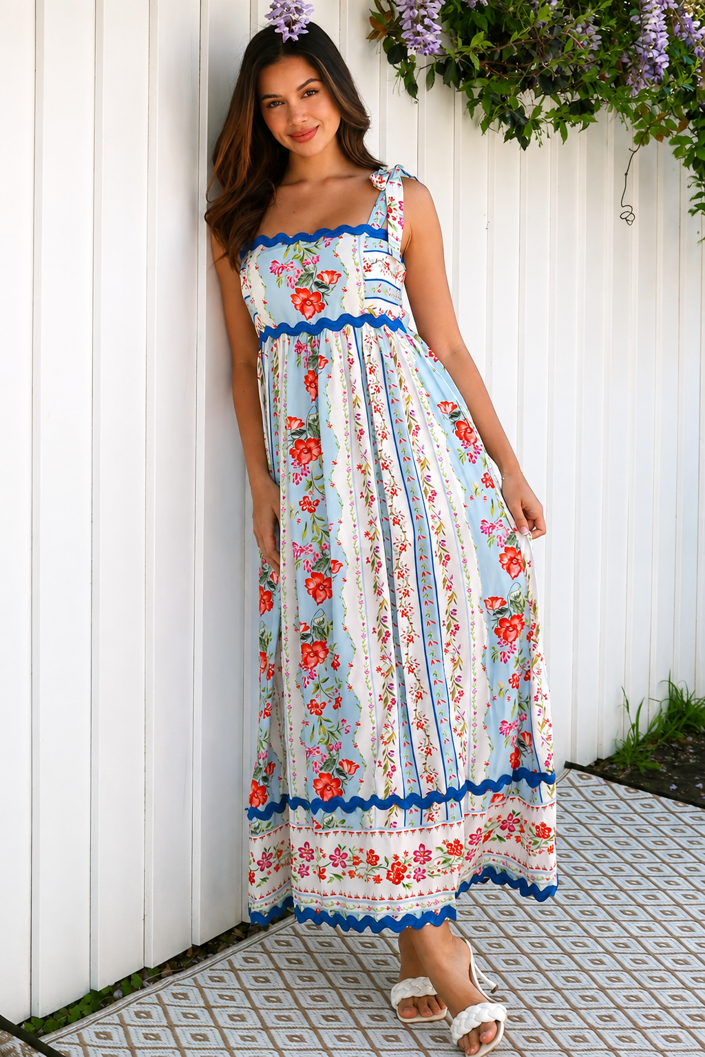 A woman wearing a sleeveless, A-line maxi dress with a white base, blue scalloped trim, and a red and pink floral print, standing outdoors in front of green plants.