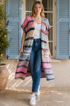 A woman wearing a long, open-front, short-sleeved chunky knit cardigan with horizontal multicolor stripes in pink, blue, beige, brown, and white, paired with a white tank top and blue jeans, standing in front of a window with blue shutters.
