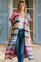 A woman wearing a long, open-front, short-sleeved chunky knit cardigan with horizontal multicolor stripes in pink, blue, beige, brown, and white, paired with a white tank top and blue jeans, standing in front of a window with blue shutters.