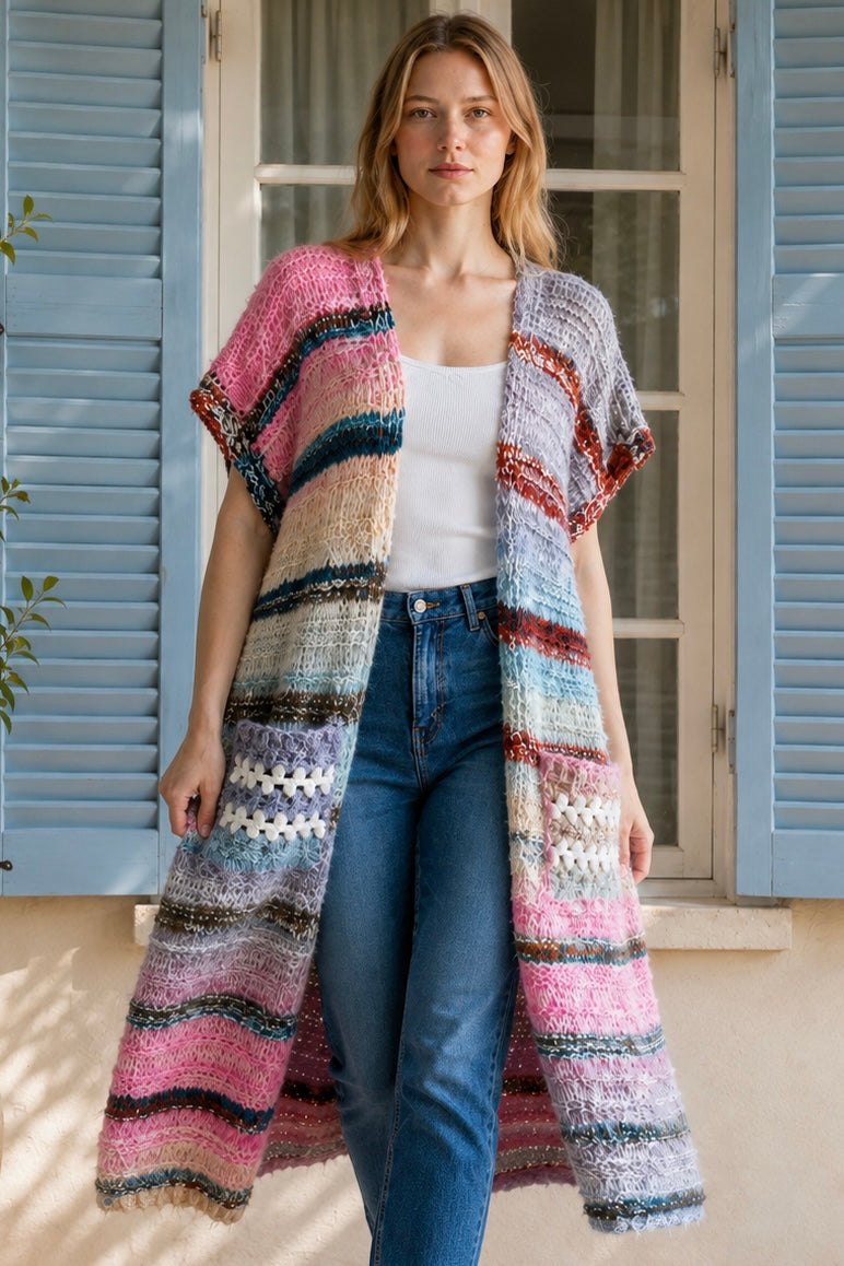 A woman wearing a long, open-front, short-sleeved chunky knit cardigan with horizontal multicolor stripes in pink, blue, beige, brown, and white, paired with a white tank top and blue jeans, standing in front of a window with blue shutters.