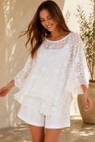 A woman wearing a white lace long-sleeve tunic top with matching white shorts, standing in a sunlit outdoor setting with a beige wall and potted plant in the background.