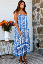 A woman modeling a blue and white patterned maxi dress with spaghetti straps, standing on a patterned tile porch next to a small table with orange flowers.