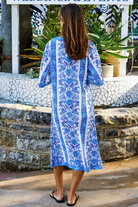 A woman wearing a blue and white patterned kaftan dress with a V-neck and three-quarter sleeves, walking outdoors in front of a stone wall and tropical plants.