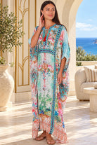 A woman modeling a long, flowing kaftan dress with a colorful floral and botanical print adorned with sparkling rhinestones standing on a sunlit outdoor patio with a view of the ocean in the background | Bohemian Love Runway 
