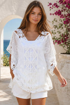 A woman wearing a white eyelet lace cover-up top with cutout details, standing outdoors in front of a white stucco wall with a blue ocean and pink flowers in the background.